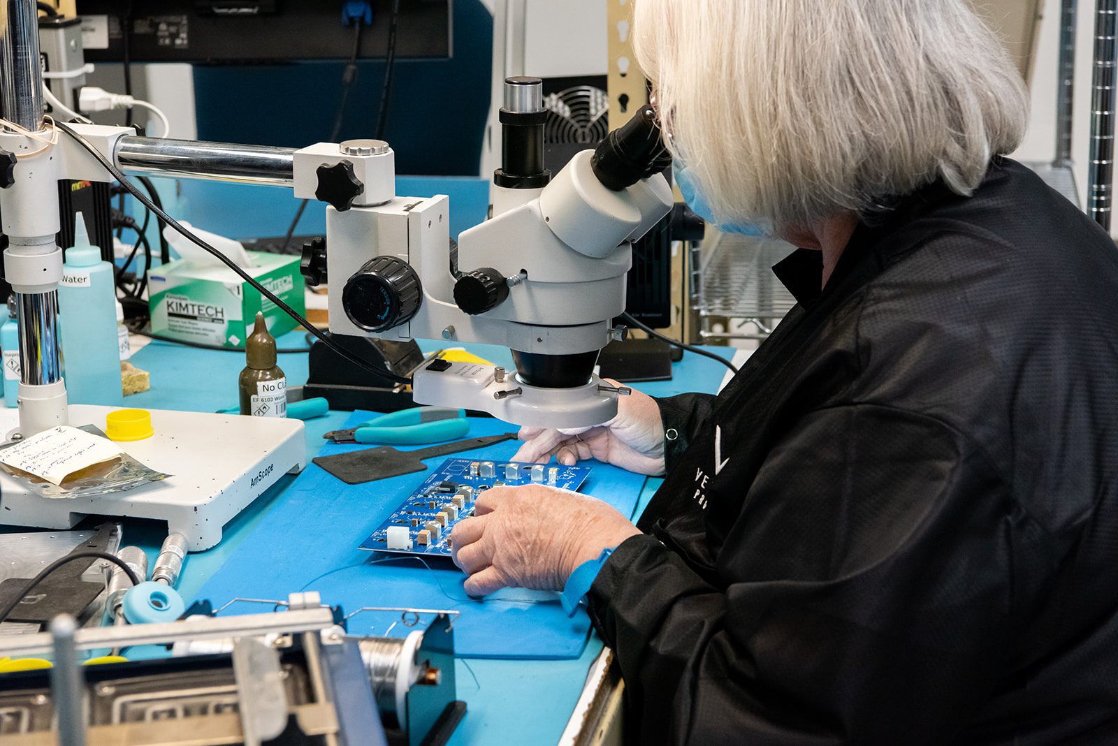 A technician in a black jacket examines a circuit board using a microscope in a high-tech lab.